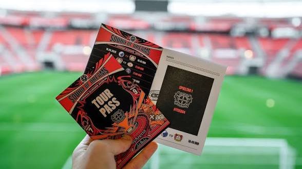 Hand hält den Champions-League-Tour-Pass im Sound-of-Dreams-Design vor dem Spielfeld der BayArena