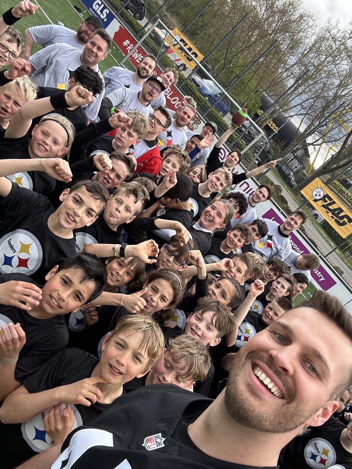 Selfie mit einer großen Gruppe jubelnder Kinder in schwarzen Steelers-Shirts auf einem Kunstrasenplatz, dahinter Betreuer und Banden