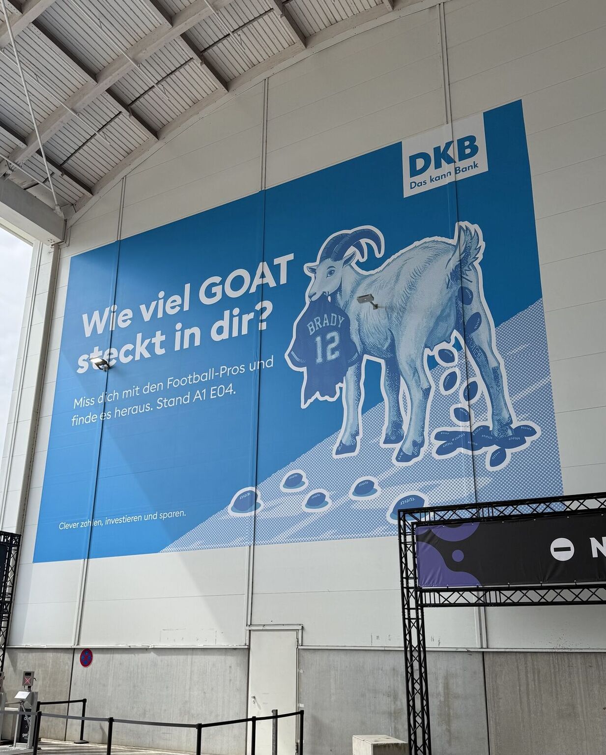 Großes DKB-Plakat an der Hallenwand: eine Ziege mit Brady-Trikot im Maul, daneben „Wie viel GOAT steckt in dir?“