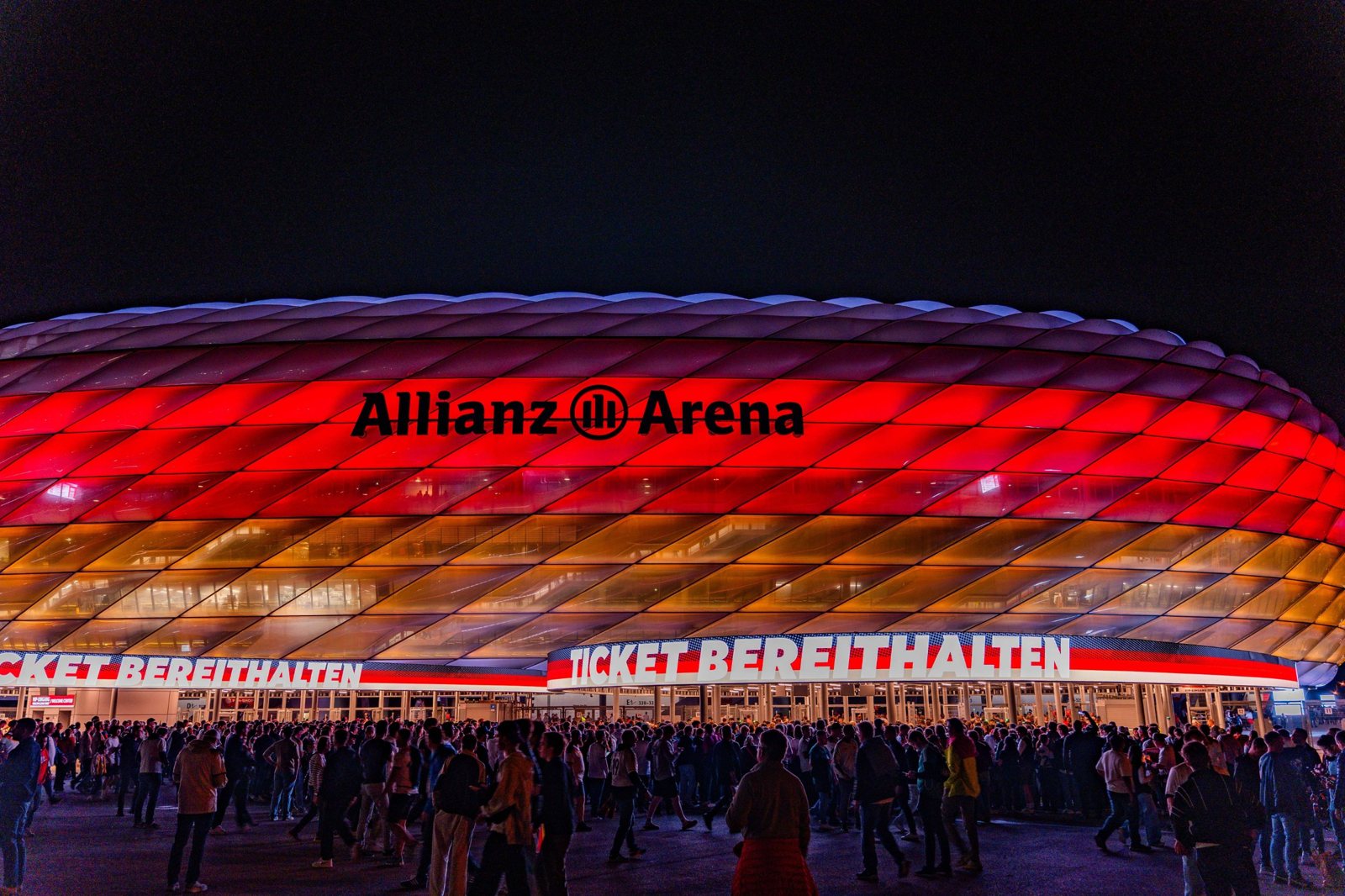 Die rot beleuchtete Allianz Arena am Abend, davor Fans auf dem Weg zum Eingang