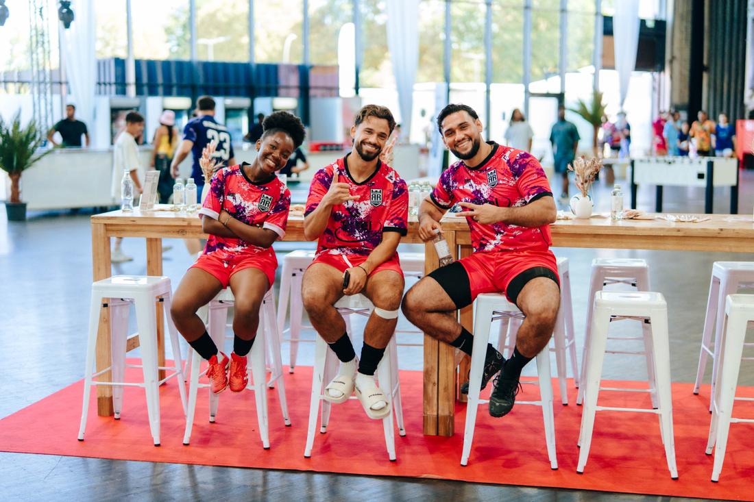 Drei lachende Spieler*innen im Royal-Roots-Trikot auf weißen Barhockern am roten Teppich, Festivalbetrieb im H