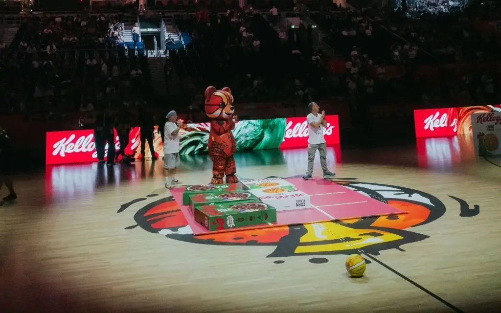 Halbzeitshow in der Arena: Maskottchen und zwei Moderatoren auf dem Court neben überdimensionalen Kellogg’s-Packungen, dahinter Kellogg’s-LED-Banden