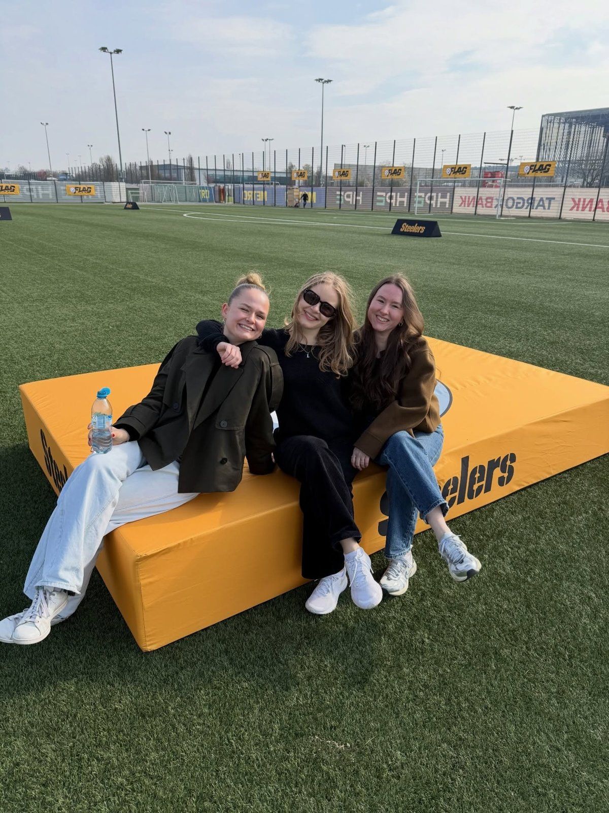Drei Frauen sitzen lachend auf einer gelben Steelers-Matte auf einem Kunstrasenplatz, dahinter Flag-Football-Banner