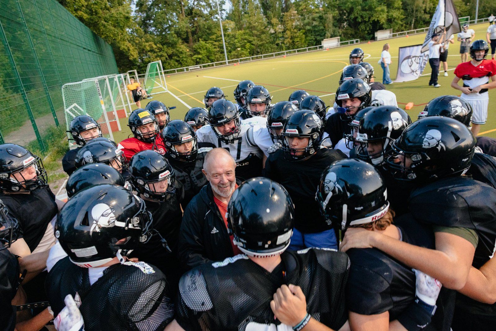 Football-Team in schwarzen Helmen im engen Huddle auf dem Kunstrasen, in der Mitte ein lachender älterer Mann in Trainingsjacke