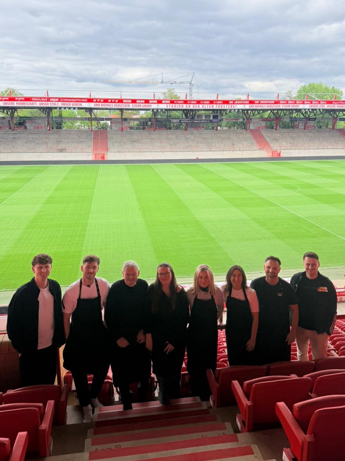 Gruppe von acht Personen, viele in schwarzen Schürzen, auf der Tribüne des leeren Stadions An der Alten Försterei