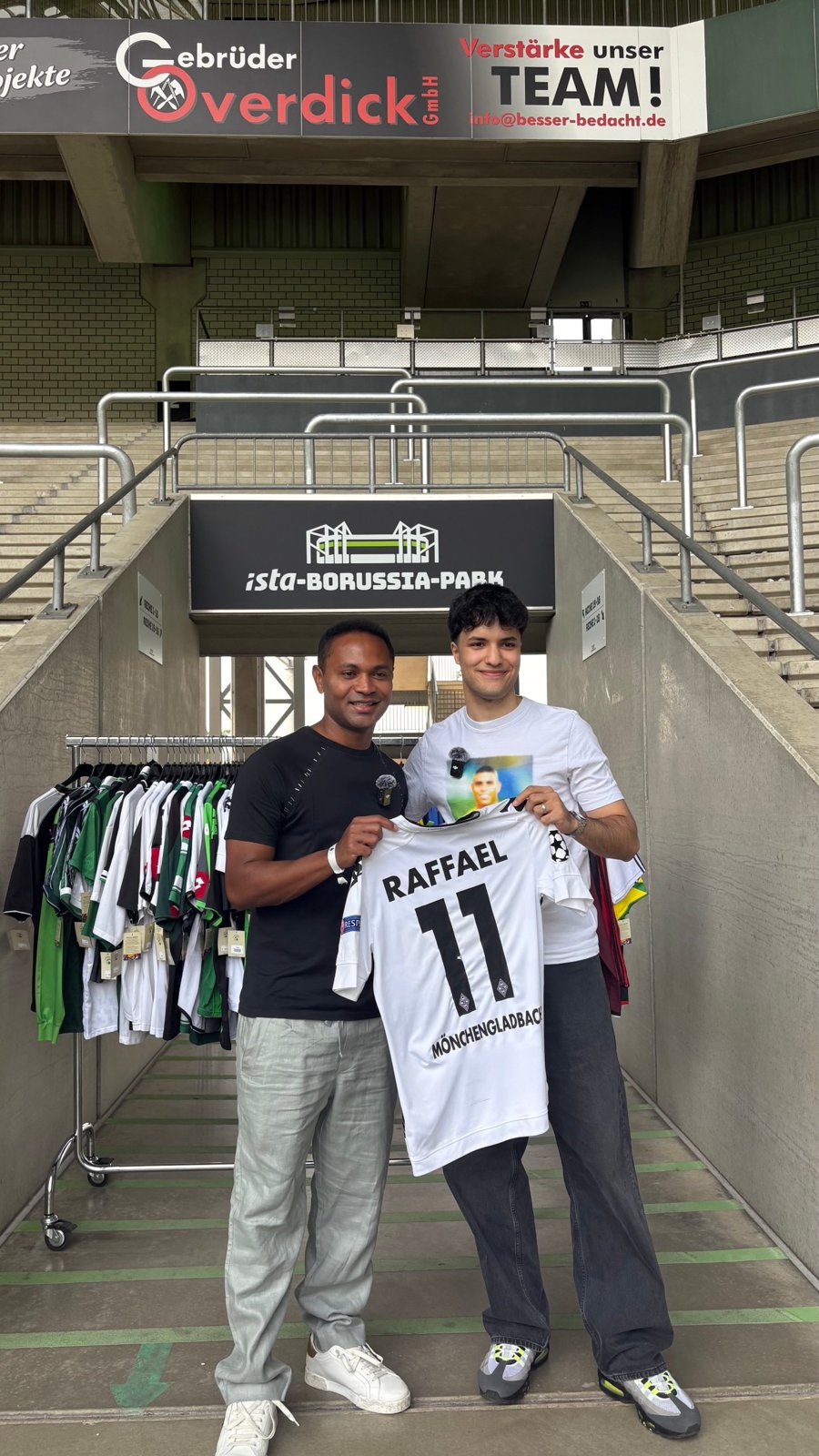 Zwei Männer halten vor dem Tribünenaufgang mit Schild ista-Borussia-Park ein weißes Gladbach-Trikot mit der Nummer 11 hoch, daneben ein Ständer mit Retro-Trikots