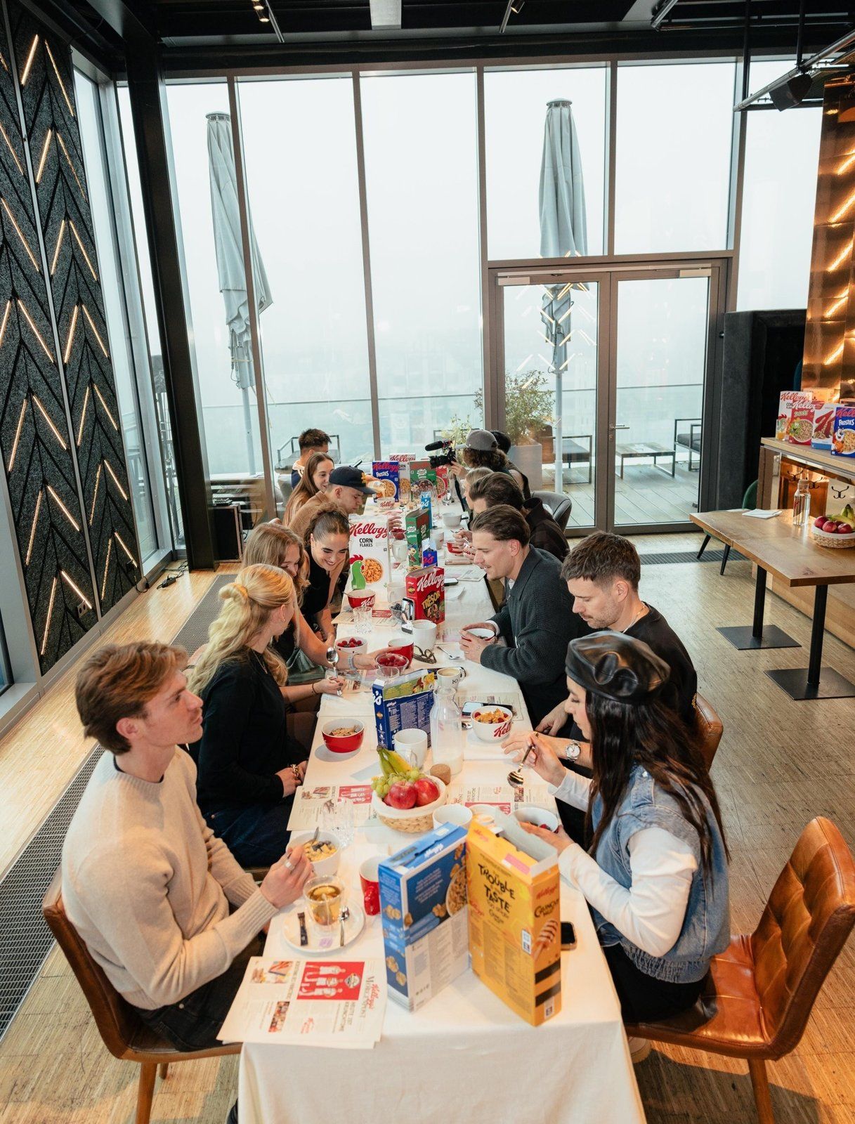 Lange Frühstückstafel mit Kellogg’s-Packungen, Schalen und Obst, an der Creator*innen vor großer Fensterfront sitzen, im Hintergrund eine Kamera