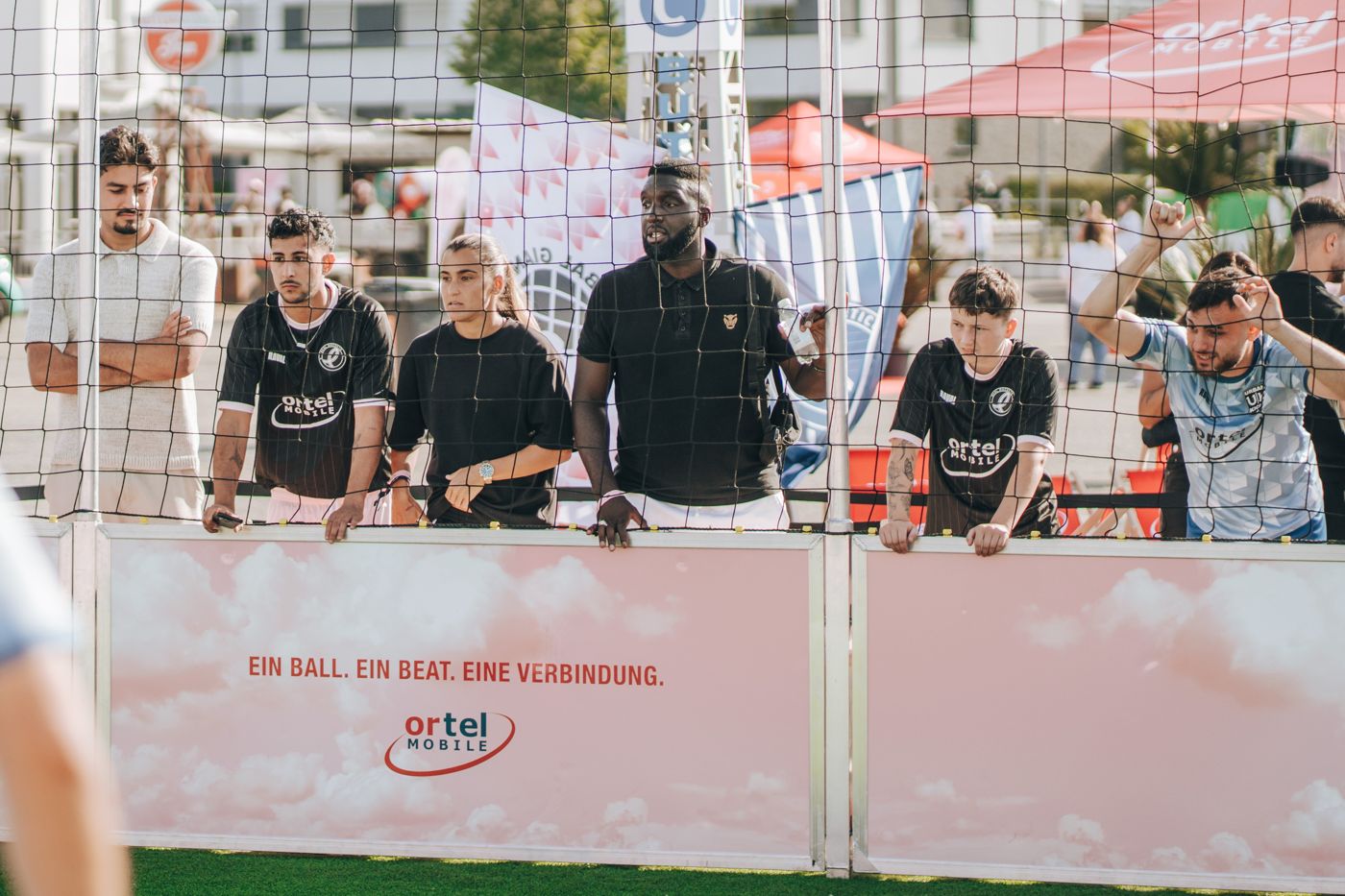 Zuschauer*innen in Trikots stehen hinter der gebrandeten Bande des Streetball-Käfigs mit Ortel-Mobile-Logo