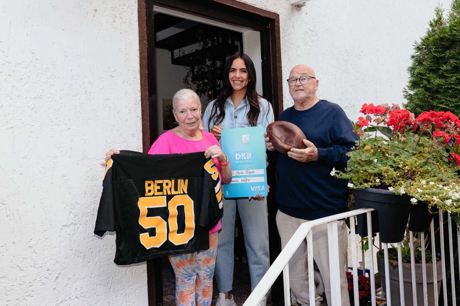 Älteres Paar vor der Haustür mit schwarzem Berlin-Trikot (Nr. 50) und altem Lederfootball, dazwischen eine Frau mit großer DKB-Ticketkarte
