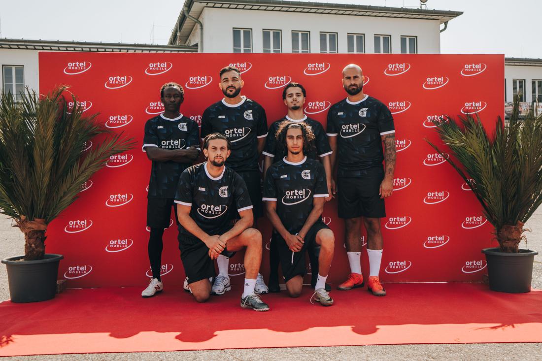Team in schwarzen Ortel-Trikots posiert selbstbewusst vor roter Ortel-Mobile-Sponsorwand mit Palmen – maximale